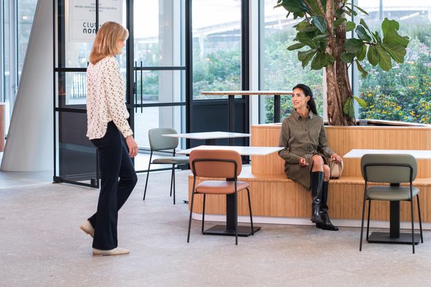 Innenansicht eines ABN AMRO Büros mit einer dunkelhaarigen Frau, welche auf einer Holzbank sitzt und eine andere blonde Frau anschaut, welche gerade ins Bild gelaufen ist.
