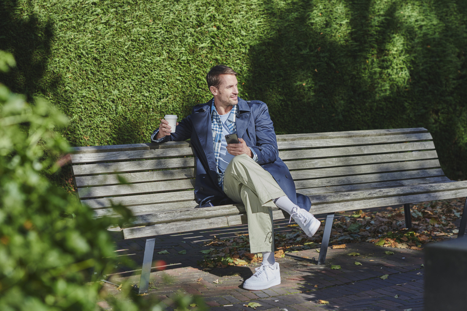 Man buiten op een bankje met een beker en zijn telefoon in zijn handen