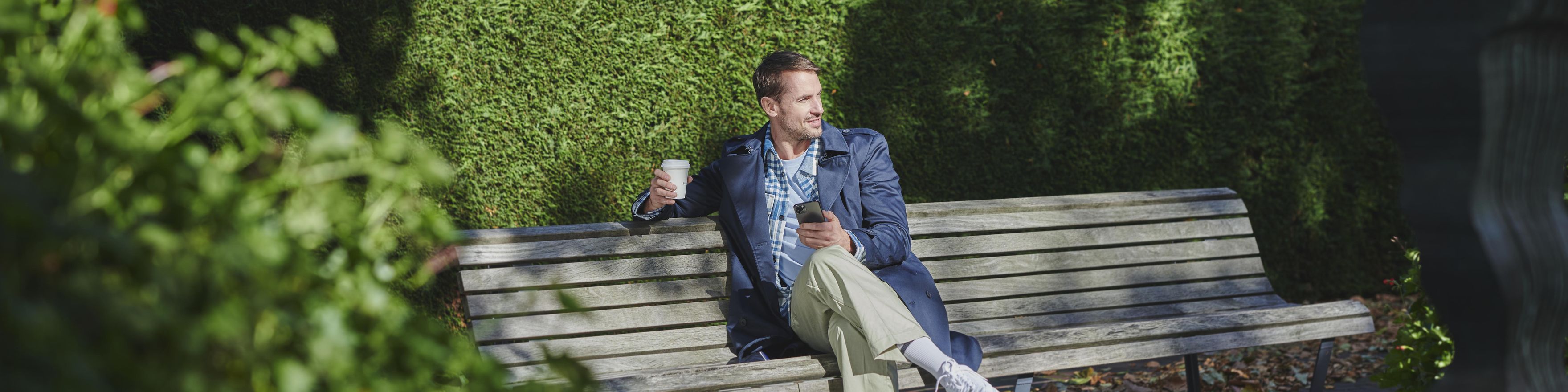 Man buiten op een bankje met een beker en zijn telefoon in zijn handen