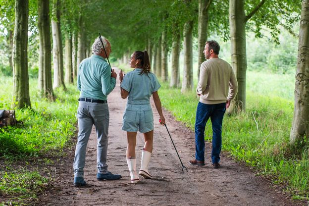 Drei Menschen stehen im Wald und laufen einen Weg entlang. Zwei von diesen Menschen sammeln Müll auf mit Müllzangen.