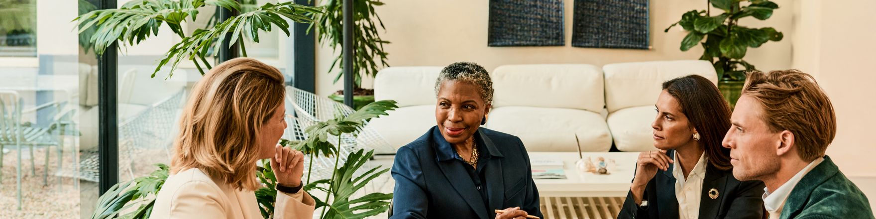 Mehrere Bethmann HAL Kunden sitzen mit einer Kundenberaterin an einem Holztisch in einem Besprechungsbereich mit großen Pflanzen, Büchern und Unterlagen. Auf dem Tisch liegen Notizbücher, Dokumente und Wassergläser.