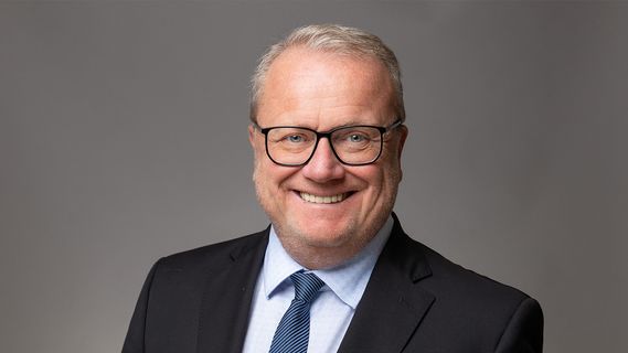 Porträt von Michael Di Martino (Niederlassungsleiter Bethmann HAL Köln) mit Brille, dunklem Anzug und blauer Krawatte vor grauem Studiohintergrund. Die Person blickt direkt in die Kamera und ist als Business-Porträt aufgenommen.