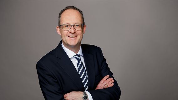 Porträt von Thomas Jäger (Niederlassungsleiter Bethmann HAL München) mit Brille in dunklem Anzug und gestreifter Krawatte vor grauem Hintergrund. Die Person steht mit verschränkten Armen und ist im linken Bildbereich positioniert, während rechts freier Raum sichtbar ist.