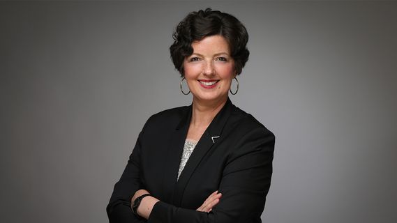 Porträt von Katja Dreps (Niederlassungsleiterin Bethmann HAL Münster) in dunklem Blazer und hellem Oberteil vor einer goldfarbenen, geometrisch gemusterten Hintergrundwand. Die Person ist leicht seitlich zur Kamera positioniert und als Business-Porträt aufgenommen.