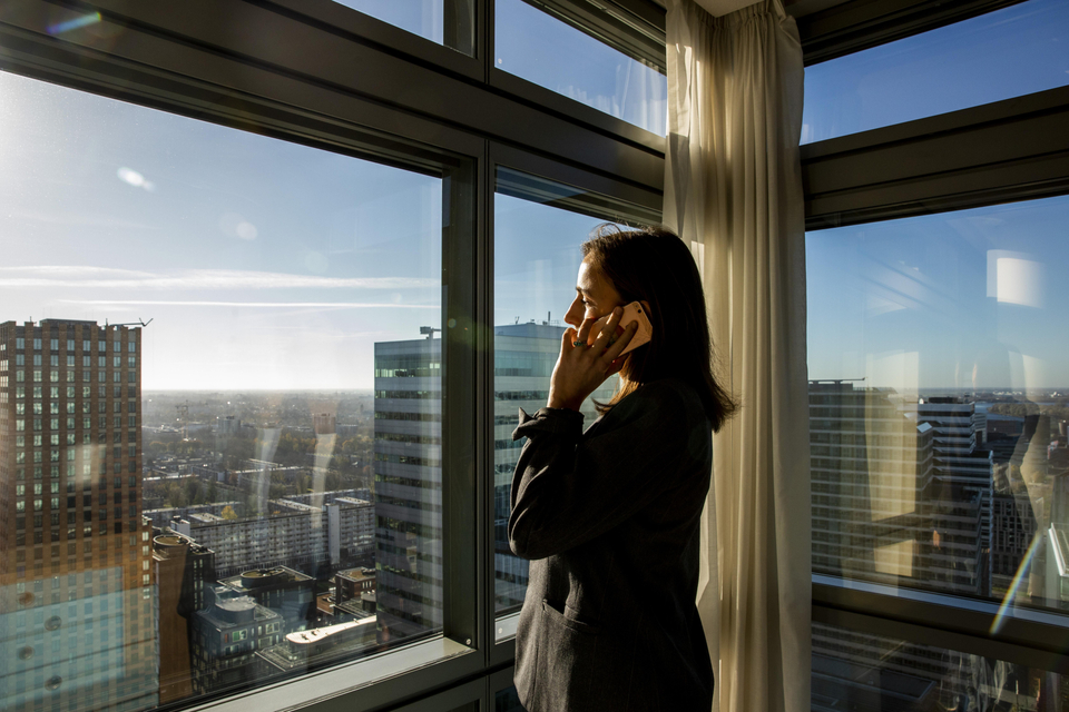 Eine ABN AMRO Bankerin mit dunklen langen Haaren und Blazer steht an einem Fenster in einem Hochhaus und telefoniert mit dem Smartphone am Ohr.
