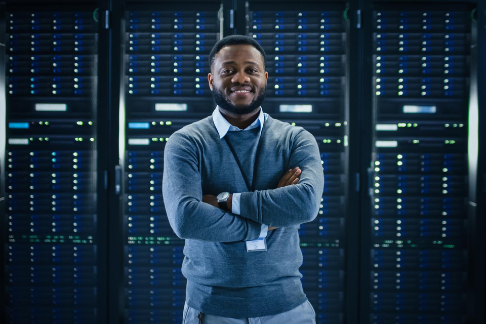 Ein junger ABN AMRO Mitarbeiter aus dem Bereich Digital Assets steht mit verschränkten Armen lächelnd vor einer Server-Wand und schaut in die Kamera.