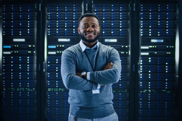 Ein junger ABN AMRO Mitarbeiter aus dem Bereich Digital Assets steht mit verschränkten Armen lächelnd vor einer Server-Wand und schaut in die Kamera.
