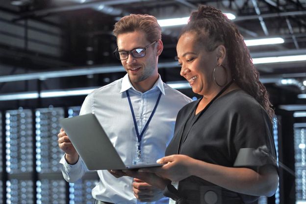 Zwei junge ABN AMRO Mitarbeiter aus dem Bereich Digital Assets stehen in einer Server-Halle und schauen gemeinsam fröhlich auf einen Computer, den die Mitarbeiterin in der Hand hält.