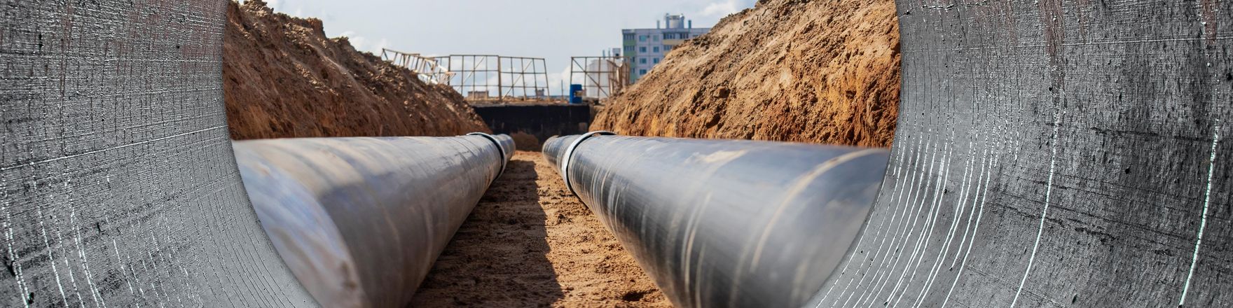 Bethmann HAL Active Investor (Im Fokus): Blick durch ein Betonrohr auf zwei parallel verlaufende Rohrleitungen in einer Baugrube. Die Aufnahme zeigt die verlegten Leitungen zwischen Erdwällen unter blauem Himmel mit vereinzelten Wolken.