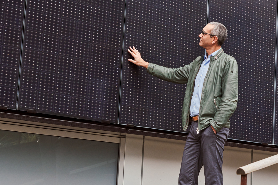 Ein ABN AMRO Kunde steht in lockerer Kleidung auf einer Steintreppe im Außenbereich und fährt mit seiner Hand über eine Photovoltaikanlage an der Wand während er seinen Blick auf sie richtet.
