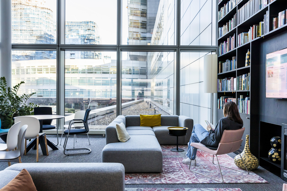 Eine ABN AMRO Kundin sitzt mit dem Rücken zur Kamera auf einem Leder-Stuhl, in einem büroähnlichen Wohnzimmer mit einem Sofa, einer großen bodentiefen Glasfassade als Fenster und liest ein Buch.