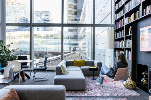 Eine ABN AMRO Kundin sitzt mit dem Rücken zur Kamera auf einem Leder-Stuhl, in einem büroähnlichen Wohnzimmer mit einem Sofa, einer großen bodentiefen Glasfassade als Fenster und liest ein Buch.